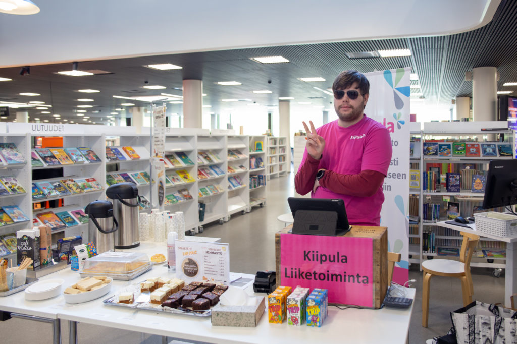 Liiketoiminnan opiskelija kahvilapisteellä Hämeenlinnan kirjastolla. Myynnissä porkkanakakkua ja omenapiirakkaa.