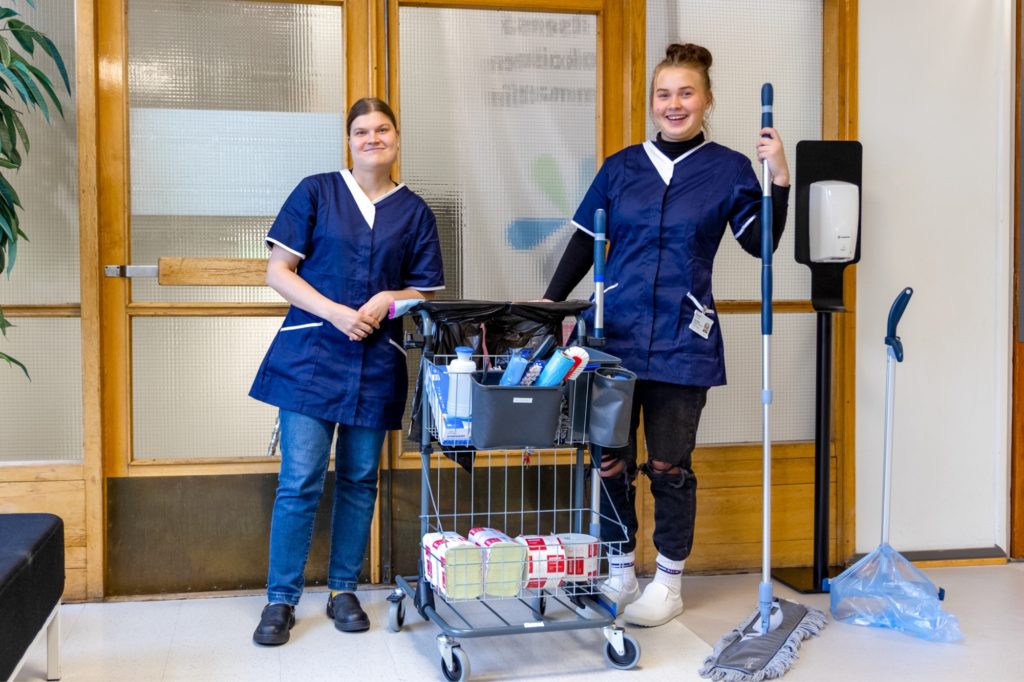 Kaksi toimitilahuoltaja opiskelijaa nojaamassa siivouskärryyn hymyillen.