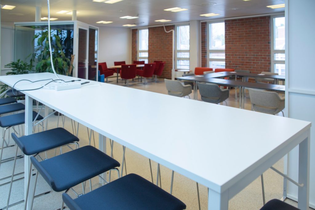 Kokoustila Maisemassa korkea kokouspöytä ja -tuolit, taustalla istumaryhmä ja kokouspöytä U-muodossa.