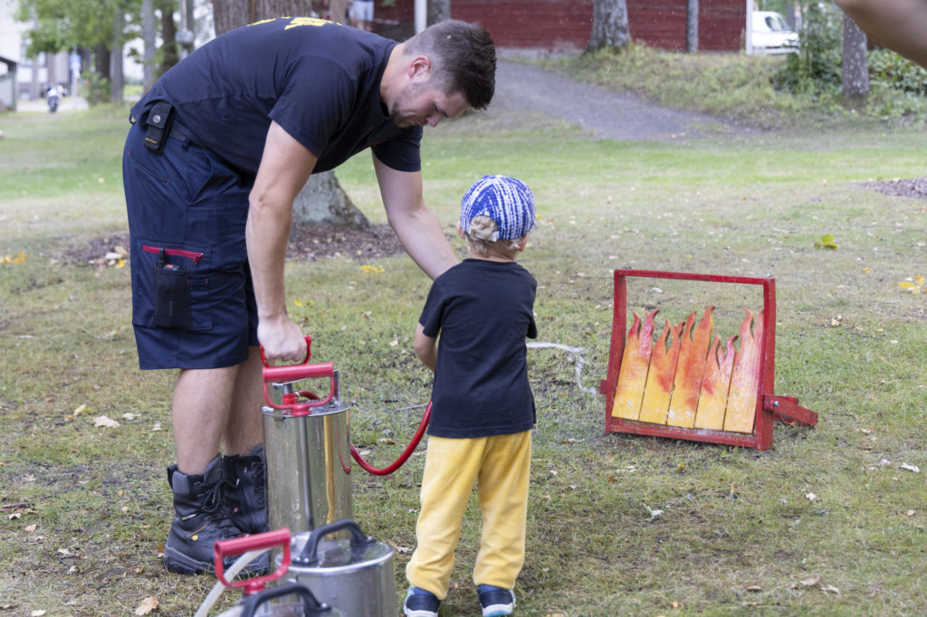 Palomies auttaa lasta sammutuslaitteen kanssa palokunnan esittelypisteellä.