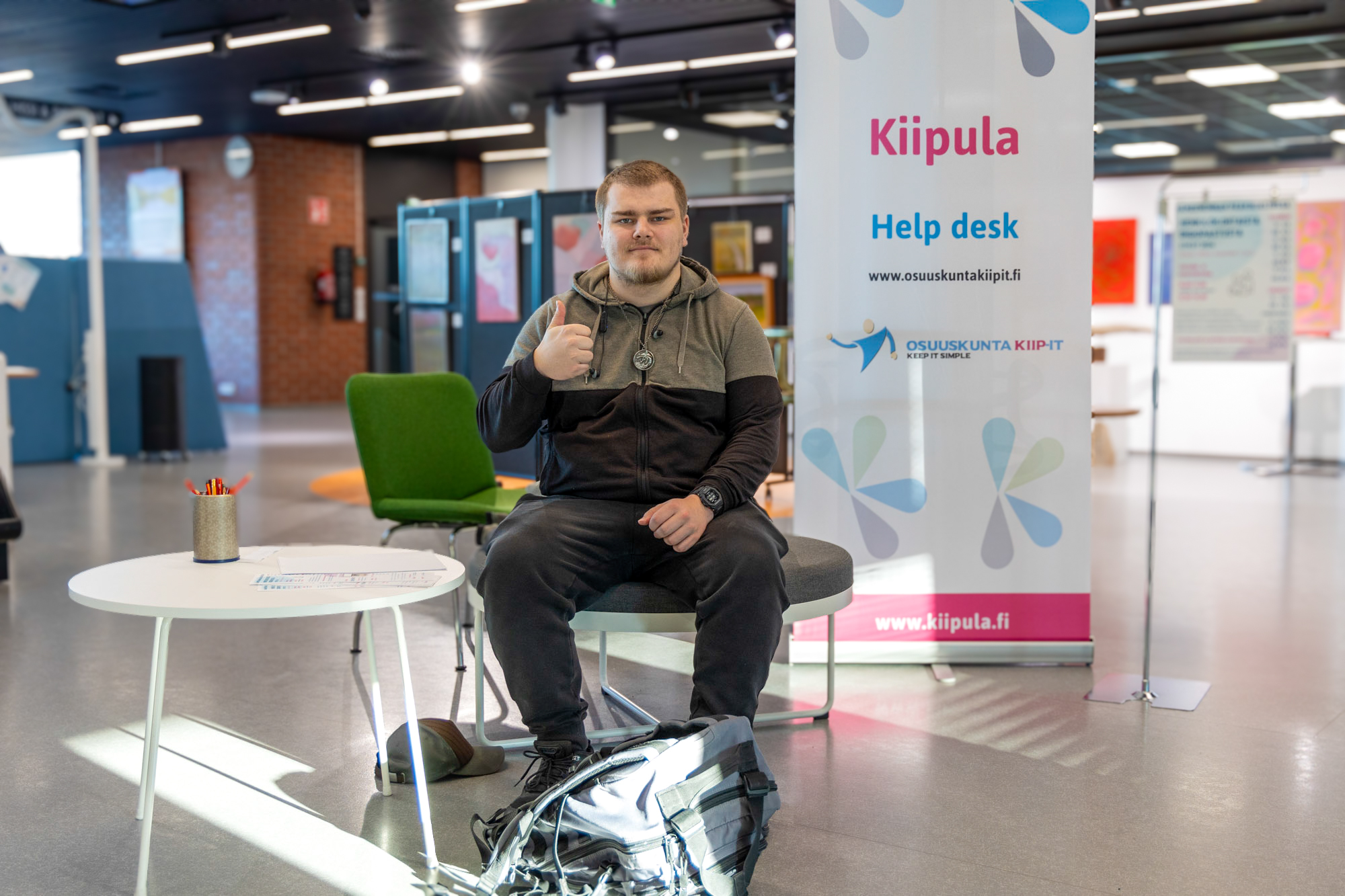 Tieto- ja viestintätekniikan opiskelija Pyry näyttää peukkua kuvaajalle. Taustalla digihelppi päivystyspiste.